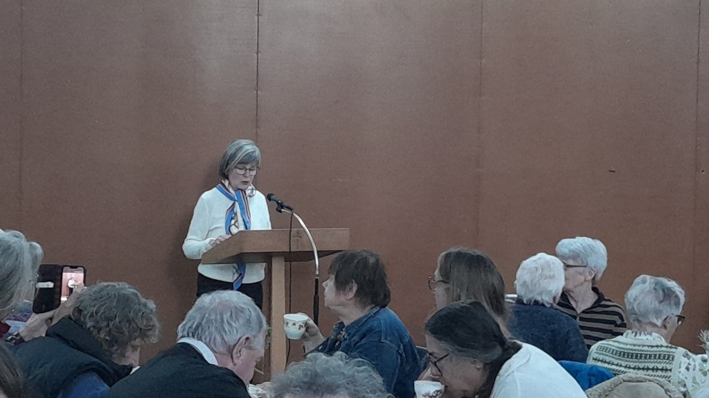A woman with short gray hair stands at a podium speaking to an audience in a community hall, with attendees seated at tables listening attentively.