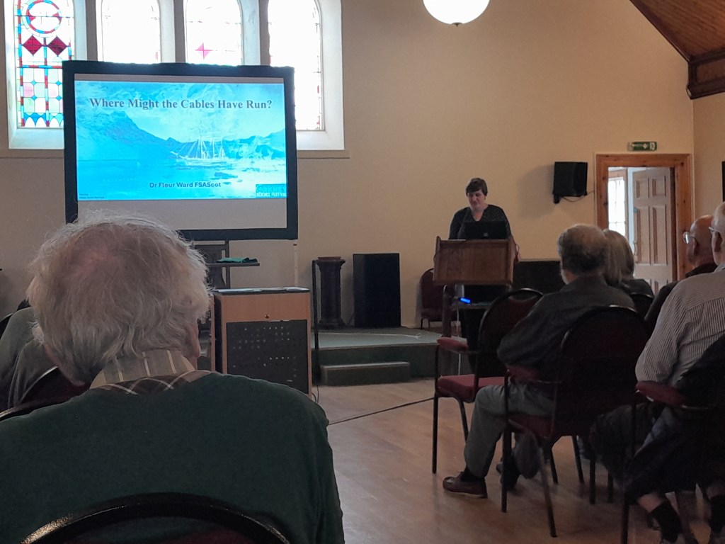 A presentation in a hall featuring Dr. Fleur Ward FSAScot, with a screen displaying the title 'Where Might the Cables Have Run?' and an audience visible in the foreground.