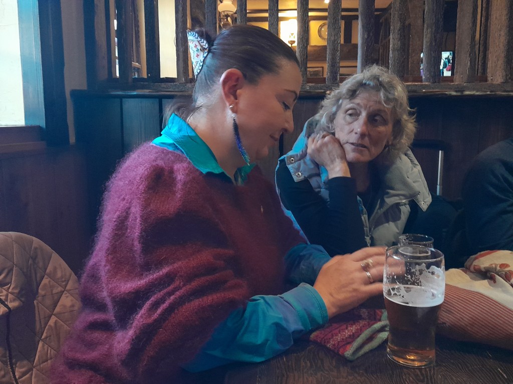 Two women sitting at a wooden table in a cozy setting, one wearing a purple sweater and a teal shirt, engaged in conversation while the other looks on thoughtfully. A glass of beer is visible on the table.