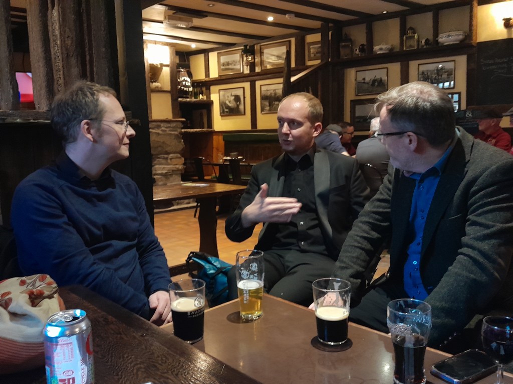 Three men engaged in conversation at a pub, seated at a table with glasses of beer in front of them, in a warm, rustic interior.