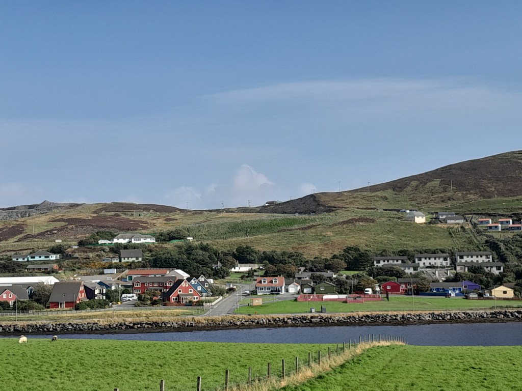 A scenic view of a coastal village with colorful houses, green hills in the background, and a calm body of water in the foreground.