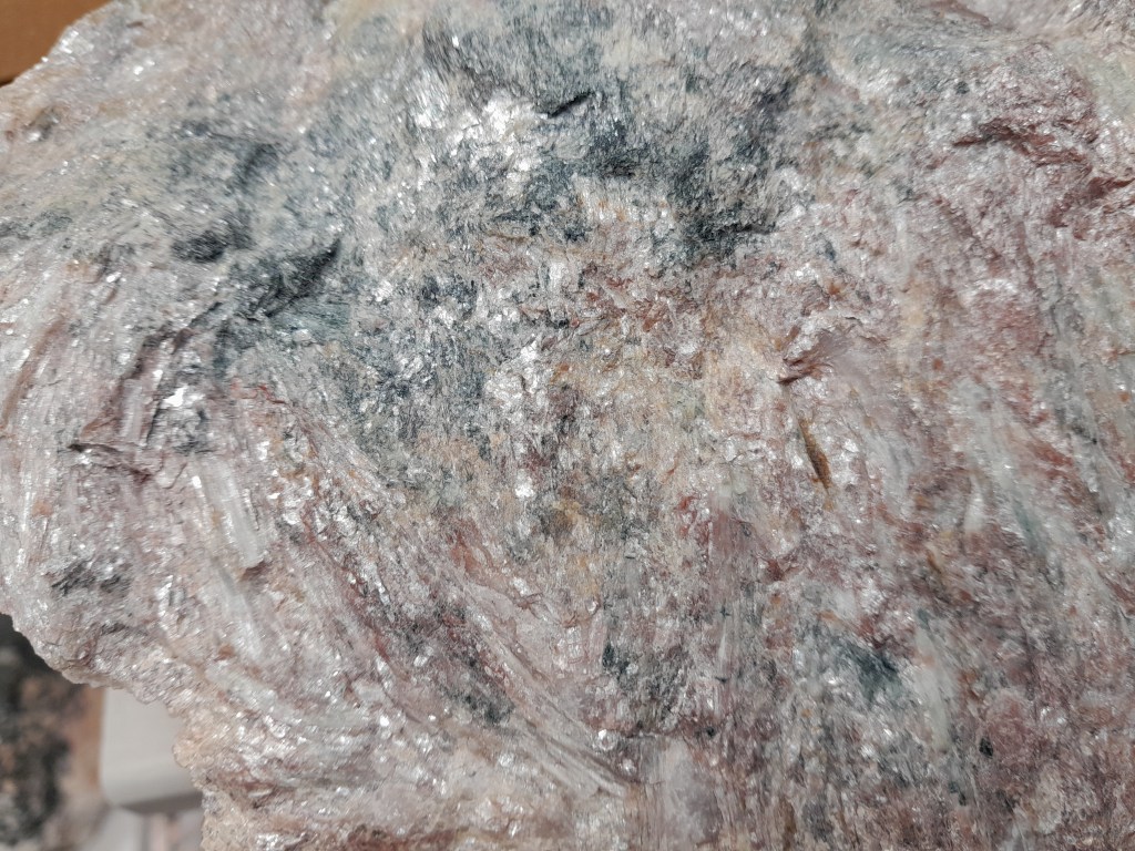 Close-up view of a mineral surface displaying shimmering, multicolored patterns with hints of pink, grey, and metallic flecks.