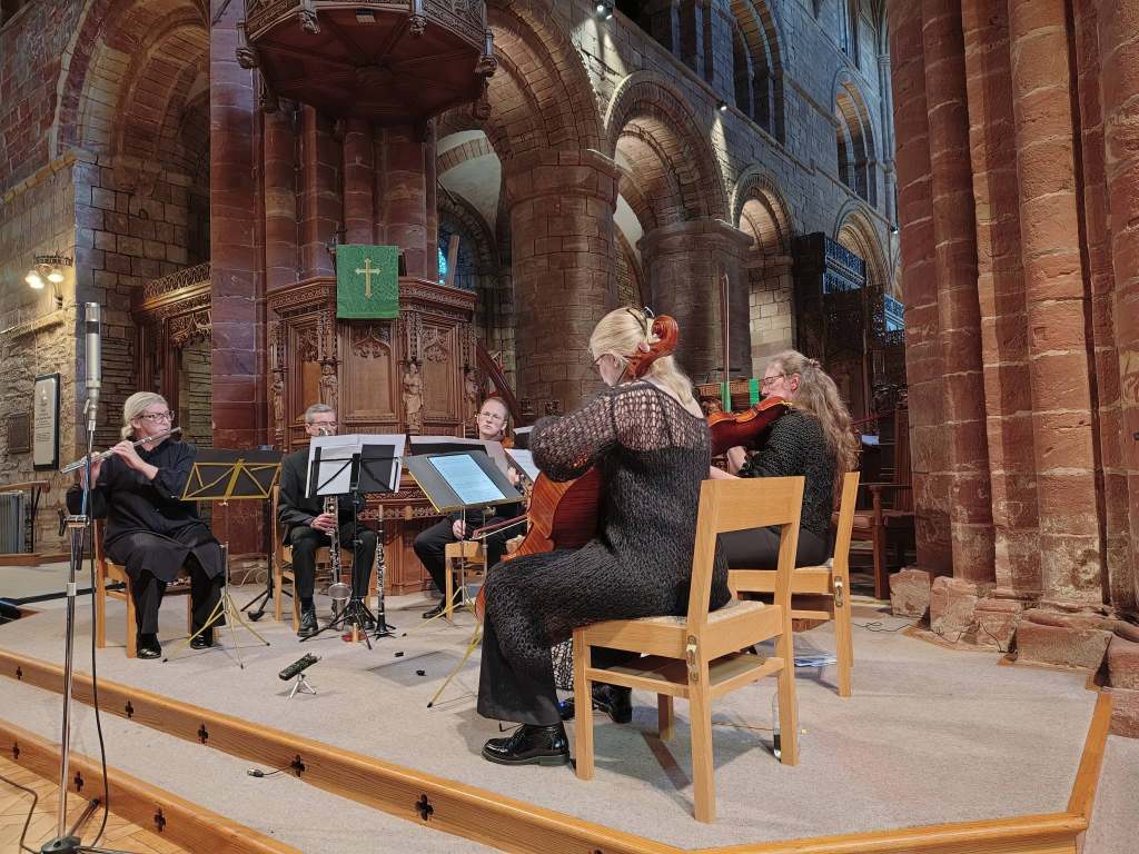 A small musical ensemble performing on stage in a church setting, featuring six musicians playing various instruments, including a flute, clarinet, cello, and violin, with wooden architecture in the background.
