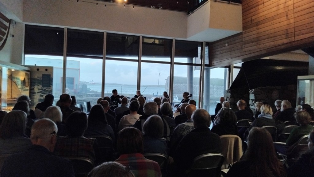 Audience seated indoors listening to a performance, with a view of a harbor through large windows.