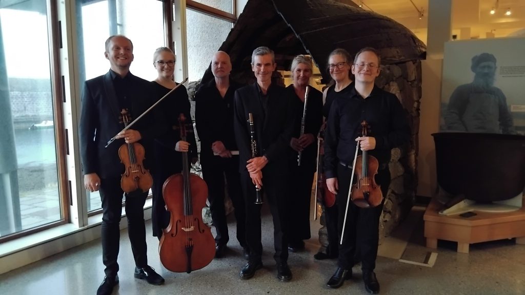 A group of musicians standing together in a museum setting, dressed in black attire. They are holding various instruments, including violins, a cello, and a flute, with a stone structure in the background.